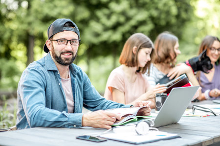 Young friends dressed casually studying with colorful books sitting at the table outdoors in the...