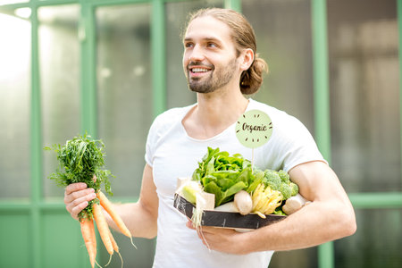 Portrait of a happy vegetarian man holding box full of fresh raw vegetables outdoors on the green backgroundの写真素材