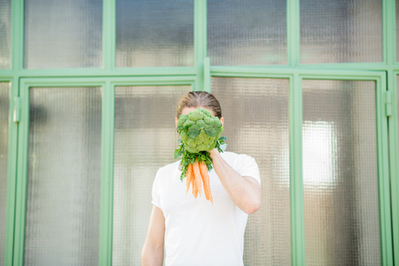 Funny portrait of a man holding broccoli with carrot instead of his head outdoors. Healthy eating conceptの写真素材