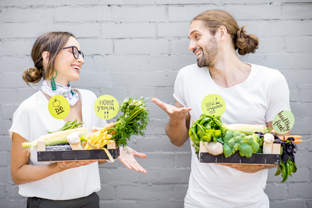 Young vegetarian couple standing with boxes full of fresh vegetables outdoors on the gray wall backgroundの写真素材