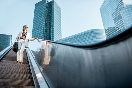 Stylish businesswoman in white suit going down on the escalator at the business centre outdoors with skyscrapers on the background in Parisの写真素材
