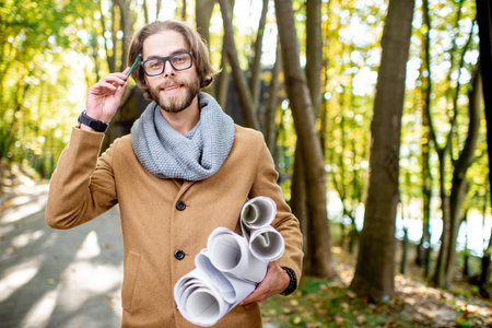 Portrait of a handsome architect dressed in coat and scarf standing with drawings outdoors in the...