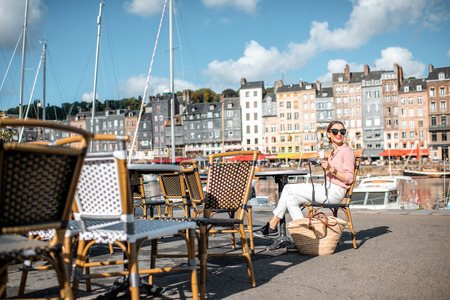 Young woman enjoying coffee sitting at the cafe outdoors near the harbour with beautiful buildings on the background in Honfleur old town, Franceの写真素材