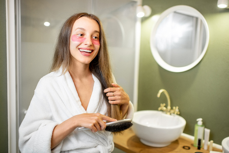 Young woman in bathrobe with patches taking care of herself combing hair in the bathroomの写真素材