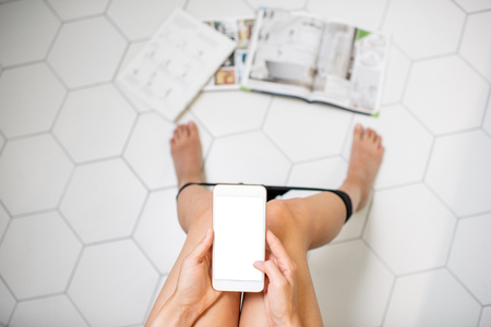 Woman holding a smartphone with white screen whilel sitting on the toilet bowl with magazines on the floorの写真素材