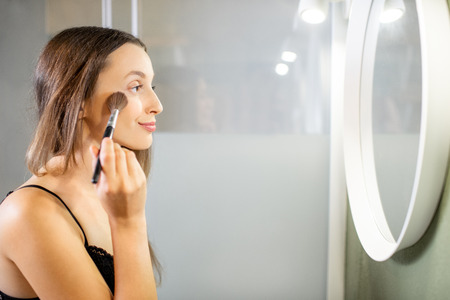 Beautiful woman in the black underwear applying makeup standing near the round mirror in the modern bathroomの写真素材