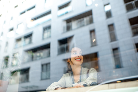 View through the window on the stylish woman sitting indoors with modern building on the reflection. Wide view with copy spaceの写真素材