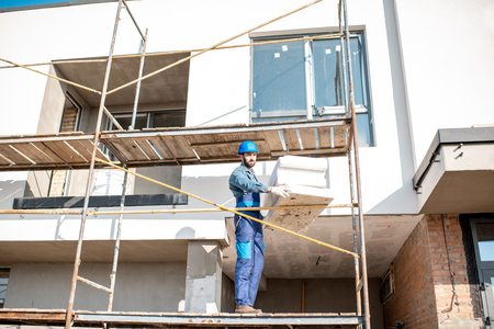 Builder warming a building facade with foam panels standing on the scaffoldings on the construction site