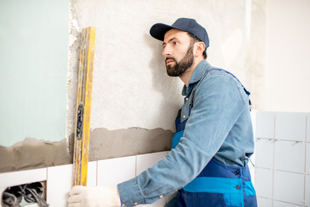 Workman in blue uniform checking the level while installing white ceramic tiles at the bathroomの写真素材