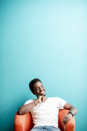 Portrait of a young african man dressed in white t-shirt and jeans sitting on the chair on the colorful background. Image with copy spaceの写真素材