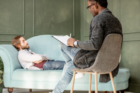 Young caucasian man visiting an african psychologist lying on the comfortable couch during psychological session in the green officeの写真素材