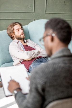 Young caucasian man visiting an african psychologist lying on the comfortable couch during psychological session in the green officeの写真素材
