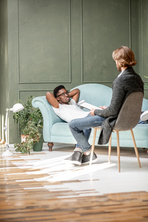 Young afro ethnicity man having a serious conversation with psychologist lying on the comfortable couch during psychological session in the office. Image with copy sopceの写真素材