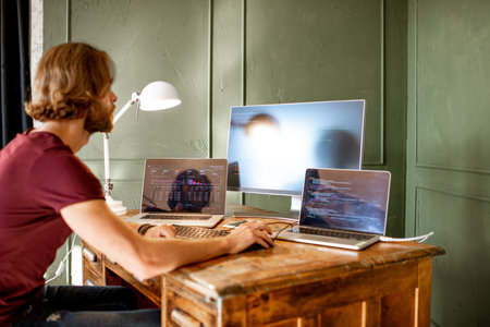 Young programmer writing a program code sitting at the workplace with three monitors in the office on the green wall backgroundの写真素材