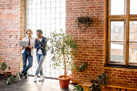 Two young businessmen dressed casually standing together near the brick wall with glass blocks in the modern officeの写真素材