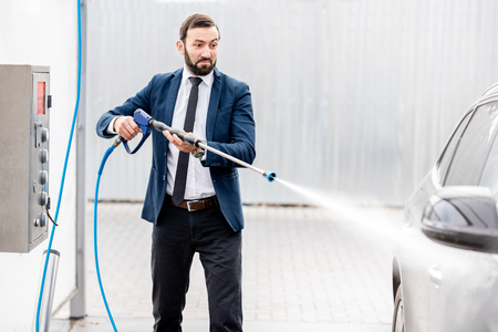 Businessman dressed in the suit washing his luxury car with washing gun on a self service open air car washの写真素材