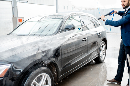 Businessman dressed in the suit washing his luxury car with washing gun on a self service open air car washの写真素材