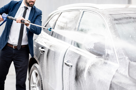 Businessman dressed in the suit washing his luxury car with washing gun on a self service open air car washの写真素材