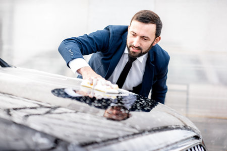 Elegant businessman dressed in the suit washing his car with yellow sponge and foam on a self...