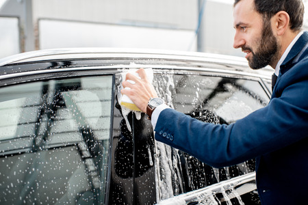 Elegant businessman dressed in the suit washing his car with yellow sponge on a self service open air car washの写真素材