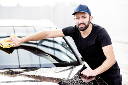 Portrait of a handsome washer in t-shirt and cap wiping windshield with yellow microfiber at the open air car washの写真素材