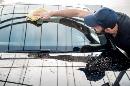 Professional washer in t-shirt and cap wiping windshield with yellow microfiber at the open air car washの写真素材
