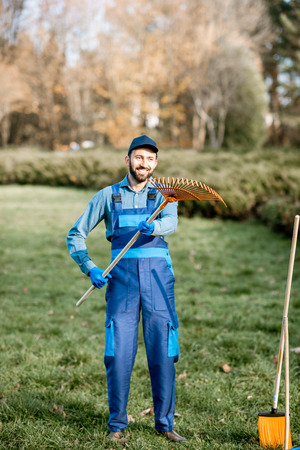 Funny portrait of a male sweeper or gardener in uniform standing with rakes in the gardenの写真素材