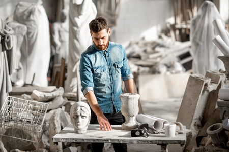 Portrait of a handsome sculptor in blue t-shirt and apron working with stone sculptures on the table at the old atmospheric studioの写真素材
