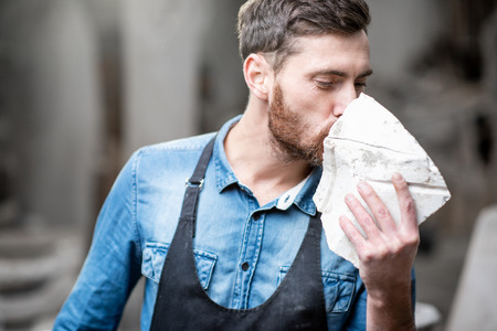 Portrait of a handsome sculptor in blue t-shirt and apron holding a piece of stone in the studioの写真素材