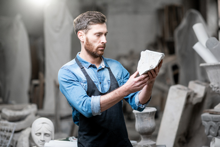 Portrait of a handsome sculptor in blue t-shirt and apron holding a piece of stone in the studioの写真素材