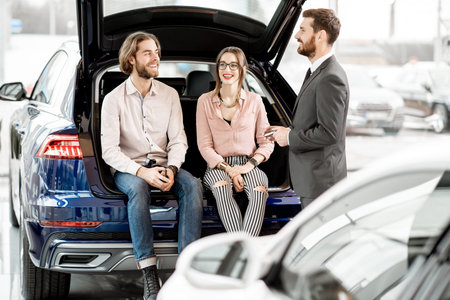 Young couple sitting in the trunk of a new car listening to the salesman at the showroom