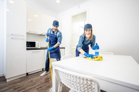 Man and woman as a professional cleaners in uniform washing floor and wiping furniture in the white...