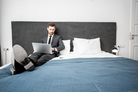Elegant businessman in the suit working with laptop on the bed of the hotel room during a business tripの写真素材