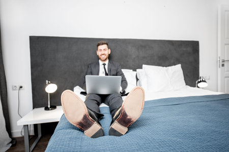 Funny portrait of a businessman in the suit working with laptop on the bed of the hotel room during...