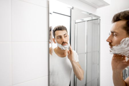 Handsome man in white t-shirt shawing his beard with blade and foam in the bathroomの写真素材