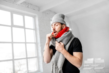 Portrait of a man wrapped in a warm scarves and hat feeling bad having a winter disease indoorsの写真素材