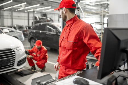 Two handsome auto mechanics in red uniform making wheel alignment with professional tools and computer at the car serviceの写真素材