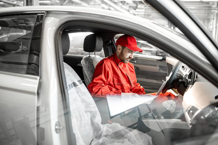 Auto mechanic in red uniform diagnosing car with computer sitting on the driver seat at the car serviceの写真素材