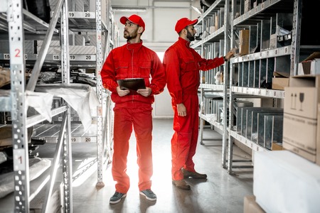 Warehouse workers in red uniform checking goods standing with clipboard in the storage with metal shelvesの写真素材