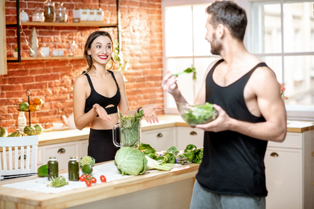 Young sports couple having snack with healthy salad and green smoothie on the kitchen at homeの写真素材