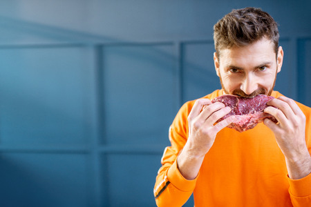 Conceptual portrait of a man dressed in bright sweater biting raw meat steak on the blue background. Image with copy spaceの写真素材