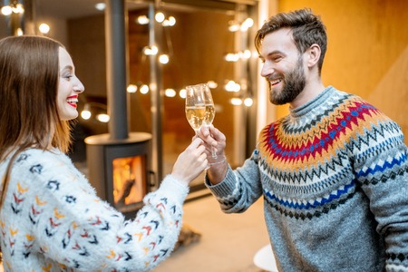 Young couple in sweaters celebrating winter holidays standing with sparkling wine in the modern house with fire placeの写真素材