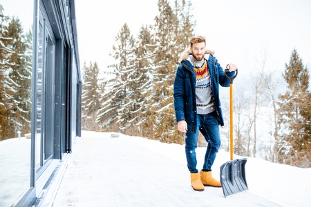 Portrait of a man in winter clothes standing with snow shovel on the terrace of the building in the mountainsの写真素材