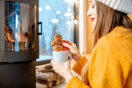 Woman holding Christmas gingerbread with cup near the fireplace at home