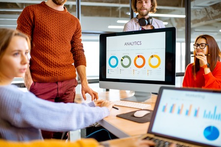 Group of young coworkers dressed casually working together on the computers with some charts sitting in the modern office interiorの写真素材