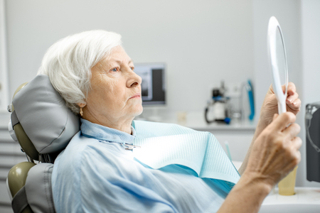 Elderly woman worried with her teeth looking on the mirror in the dental officeの写真素材