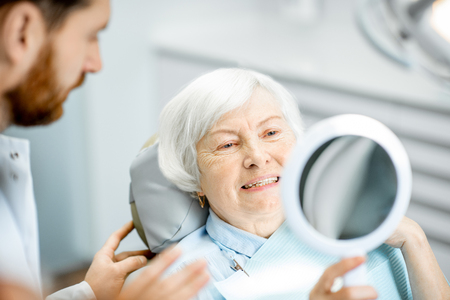 Happy elderly woman enjoying her beautiful toothy smile looking to the mirror in the dental officeの写真素材