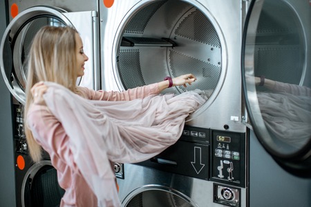 Smiling woman taking out clean and dried clothes from the drying machine in the self-service laundryの写真素材
