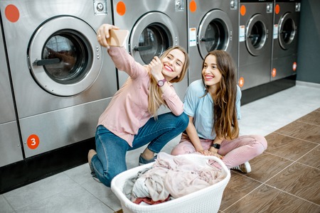 Two cheerful girlfriends making selfie photo with basket full of clothes in the professional laundryの写真素材
