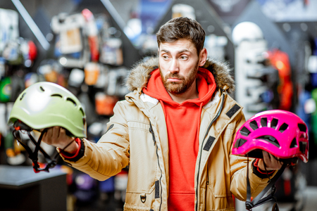 Man in winter jacket choosing mountaineer equipment holding helmets for hiking in the sports shopの写真素材